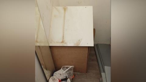 Council officers found tobacco and other items in a hole in the store covered by a wooden box with a white lid. There is a bag of rubbish next to the  box and some grey panels in front of it.
