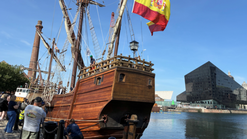 A large wooden ship is docked by Liverpool's water front. It is flying a Spanish flag and people are taking photographs