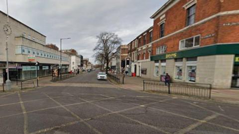 A box junction with yellow markings is at the bottom of the photo. Higher up the generic image, the road has buildings on either side of the street and there is a white car at traffic lights.