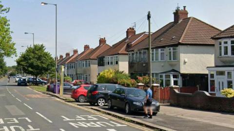 A row of semi-detached house on a dual carriageway. A number of cars are parked outside houses and a boy is about to cross the road.