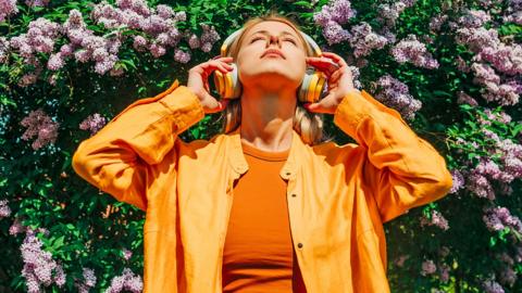 A woman wearing an orange shirt listening to music through her headphones, under a purple lilac tree outdoors.
