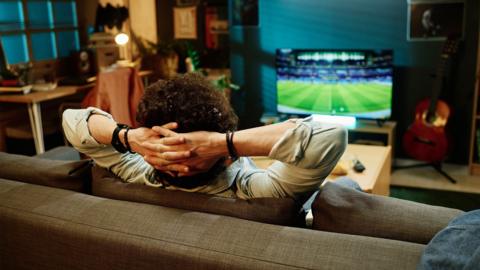 Rear view shot of man sitting relaxed in living room watching sports event on TV channel, stock image