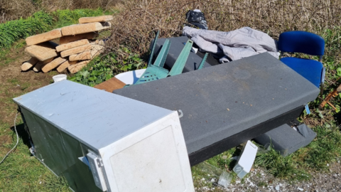 A fridge, bus seat and cushions along with other rubbish on a patch of grass.