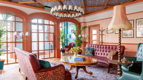 Large room with wooden windows. There are rust coloured and green sofas and chairs with flowered William Morris fabrics. There is a shiny wooden coffee table with books and Tulips on it. The walls are painted in peach and blue and there are large green plants and a chandelier along with a standard lamp and desk lamps which are wooden with cream shades. The floor is wooden with a flowered rug. The ceiling is wallpapered in blue William Morris flowered print.