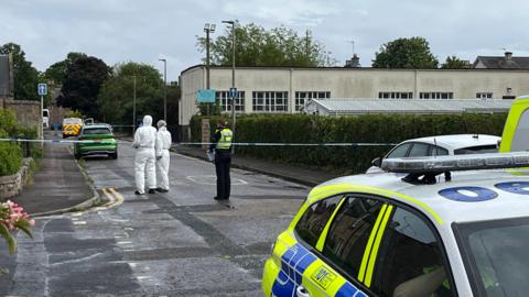 A police car is in foreground. Behind that are two people in white forensic suits, standing next to a uniformed police officer. They are behind police tape put up across a road. 