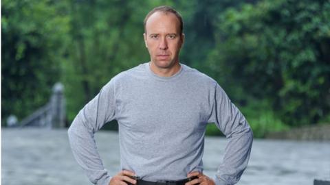 Matt Hancock wearing a grey t-shirt and green army cargo pants. He is sstanding in front of a river with his hands on his hips.
