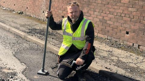 A man in a high-visibility yellow vest and dark work clothes crouches on a residential road, holding a long-handled tamping tool in one hand and a smaller tool in the other. Behind him is a red brick wall with a grass verge above it, and loose gravel and debris are visible on the road surface. He has sunglasses pushed up on his head and a short beard, and is looking directly at the camera.