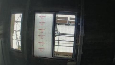 A dim, unfinished room with exposed brick walls and a row of window openings, with scaffolding visible outside.