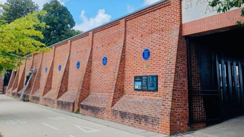 A red bricked-wall with blue plaques lined up on it. There is a person in the background on a ladder putting up another plaque.