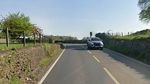 A single-carriageway road with stone walls and fields on either side and a single grey vehicle - a truck - driving towards the camera