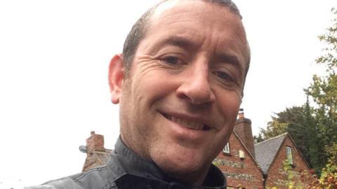 A selfie taken by David Carrick, who has short dark hair. The black collars of his jacket are pictured and a house is behind him. He is smiling.
