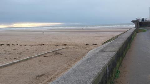 Starr Gate beach in Blackpool with the tide out and two people in the distance walking on the sand heading towards the waves and two people looking out stood behind railings on the promenade