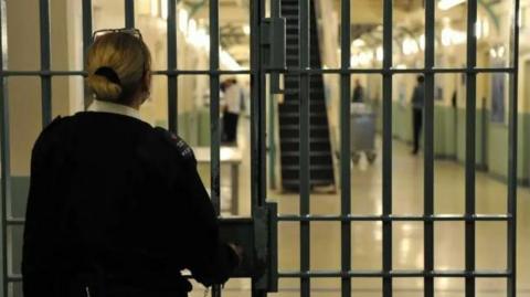 A prison goaurd with her hair tied back faces away from the camera and looks through a door made up of metal bars into a jail wing area beyond, She appears to be using a key in a lock in the door at waist height.