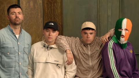 Four young men are standing in a room looking at the camera. The man on the left is holding his hands behind his back and wearing a denim top with a white top under it. The two men next to him are wearing caps on their heads and the fourth man is wearing a green, white and orange balaclava over his face.