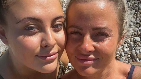 Close up selfie of Danielle and Chloe at a pebbled beach. Chloe, on the left, has blonde hair. Danielle, on the right, also has blonde hair. Both are smiling looking at the camera with their one cheek touching. 