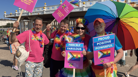 Antonois Bakiuris, Charley Ashbury, Rachel Brett and Paul Hilly stand in front of Hove Lawns ahead of the Brighton Pride parade in 2025. They hold placards depicting the Intersex-Inclusive Progress Pride flag with an added red ribbon on it, an international symbol for HIV awareness and support for people living with HIV. The signs also read "fiercely fabulous" and "proud, positive, powerful," promoting the work and stance of The Sussex Beacon. They are dressed in pink branded t-shirts while wearing rainbow face paint, glitter and leis. Paul Hilly is also holding a rainbow umbrella.