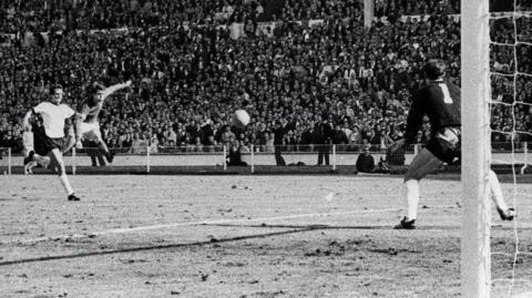 Black and white image shows England's Geoff Hurst in mid-air with his left arm and left leg outstretched as he cracks a shot past German goalkeeper Hans Tilkowski to score the final goal of the World Cup Final against West Germany at Wembley in 1966.