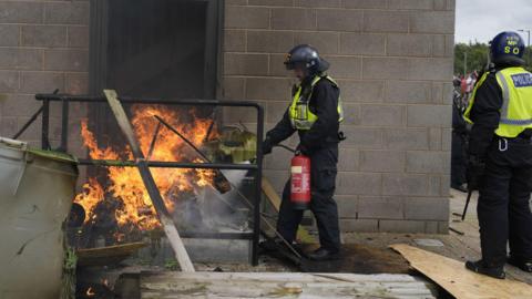 A fire is extinguished by police officers outside a door. Several planks of wood and other items can be seen engulfed by flames.