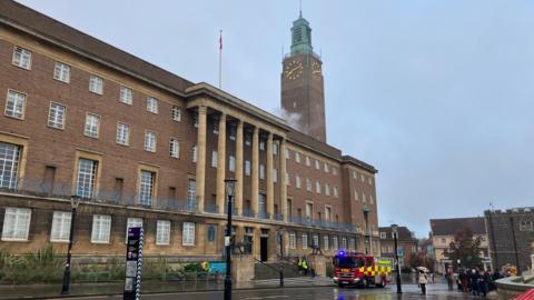 Wisps of smoke can be seen near a clock tower on the roof of City Hall in Norwich. It is an imposing civic building that is four storeys high. It has six columns above a flight of steps leading to the entrance. A fire engine is parked on the rain-soaked street outside.