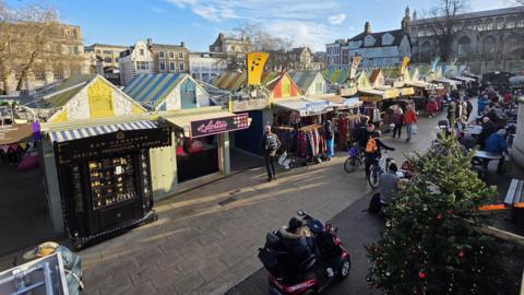 Part of Norwich Market, featuring a number of multi-coloured, striped stalls. It's quite busy with a number of shoppers in the foreground and a Christmas tree.