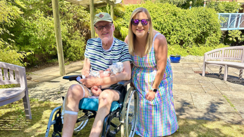 A man with a striped tshirt, shorts and cap in a wheelchair holding a baby next to a woman with long blonde hair, sunglasses and a long patterened dress in a sunny garden