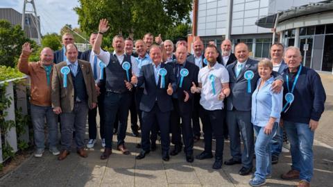 A group of Reform politicians wearing light blue rosettes and smiling.