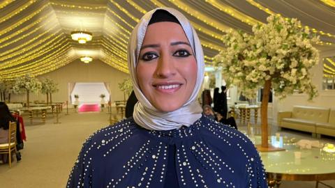 A young woman smiles at the camera while standing in the marquee, which has fairy lights strung across the ceilings. The room is set up as if for a wedding with round tables, white blossom trees and gold chairs. She wears a silver headscarf and blue blouse with sequins on, and has a small nose ring.