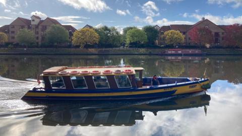 The blue and yellow ferry can be seen gliding through the harbour. The harbourside can be seen behind her, with the trees green, yellow and red behind her. 