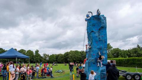 A large blue climbing tower with various young people attempting to climb it in front of a small crowd under a blue gazebo with other children waiting their turn