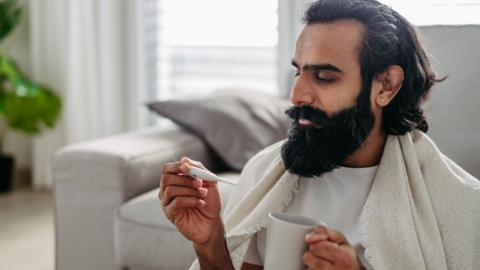 A man with a blanket over his shoulders holds a thermometer and a cup. He is sat down and leaning against a sofa.