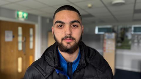 Kyran looking at the camera wearing a blue track top and a black coat on top. He has very close-cropped, black hair and a beard and moustache.