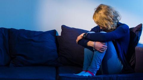 A woman with curly blonde hair sat on a navy blue sofa. She has her head down and arms around her legs.