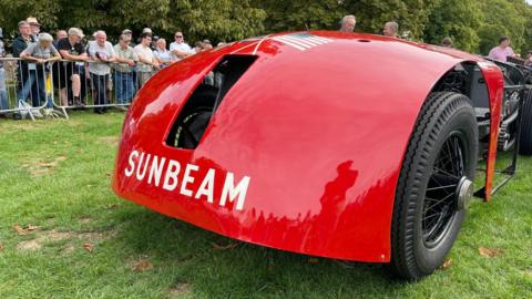 Sunbeam 1000hp in front of spectators at the National Motor Museum. The large wheels and red-painted front of the car are in the foreground, with an exposed engine to the rear.