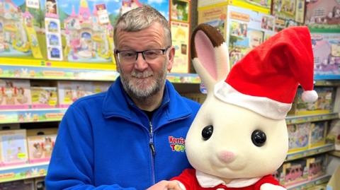 Steve Kerrison stood inside a toy shop with toys in the background. he's wearing a blue fleece jacket and holding a large bunny like toy with a Christmas hat