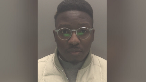 The mugshot of a man wearing transparent glasses and white jacket over a grey jumper. 