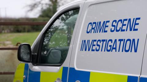 A close-up of a West Yorkshire Police van. It is mainly white, with the words "Crime Scene Investigation" written on the side in blue. There is a gate and a field in the background.