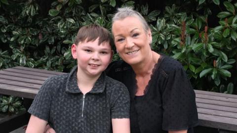 Tony Hudgell stands next to his mum Paula, who is sat at a wooden picnic bench. They are smiling at the camera and holding hands.