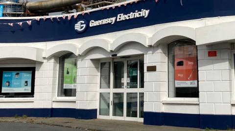 The front building of Guernsey Electricity - the sign above the glass double door, with three windows in vision. The building is painted striped blue and white.
