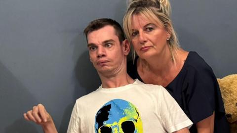 Thomas Howe, with short dark hair, wearing a white T-shirt with a blue and yellow skull design, and red trousers. He is sitting on the floor with his mother behind him. She has long blonde hair and is wearing a black top. There is a large teddy bear behind them along with some cushions. There is a grey wall in the background.