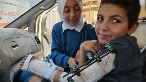 Adam al-Najjar sits in an ambulance with a brace around his arm wearing a dark t-shirt while his cousin, wearing blue and a white headscarf, tends to him.