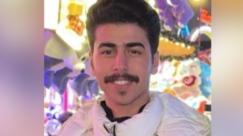 Mr Algasim is smiling at the camera while wearing a white winter coat. There are bright lights and teddies behind him, it looks like he could be at a fun fair stall. He has dark hair that is slightly quiffed at the top, as well as thick eyebrows, and a moustache.