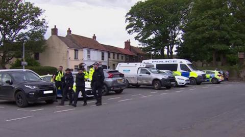 Police officers and police vehicles in a car park in Easington. Officers are wearing dark blue overalls and caps with some wearing high visibility jackets. They are huddled in a group and standing in front of a black Range Rover and a marked police car.