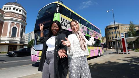 Two women pose in front of a double decker bus