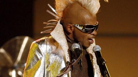  Funk music pioneer Sly Stone in a blond mohark and gold jacket, wearing sunglasses and singing into two microphones 
