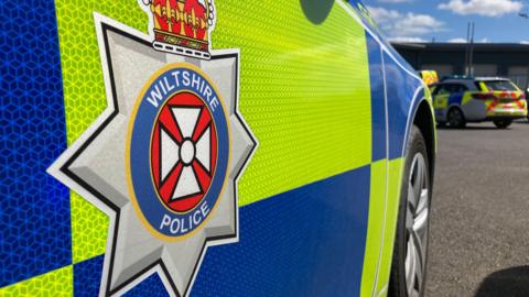 The side of a Wiltshire police car, which is parked in a car park. Another branded police car is in the background.