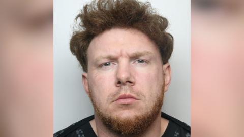 A police mugshot of Jacob Gallagher. He has wavy ginger hair and a ginger beard. He is wearing a black jumper.