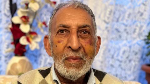 Hukum Dad with grey hair and a beard is wearing a yellow and brown jumper. He is sitting by a window