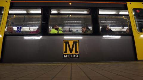 A grey, black and yellow metro train is pulled up to a platform. Passengers look out of the window.