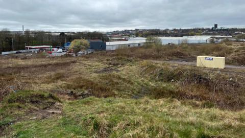 An area of land behind a set of industrial units with a petrol station and Tunstall town centre in the distance.