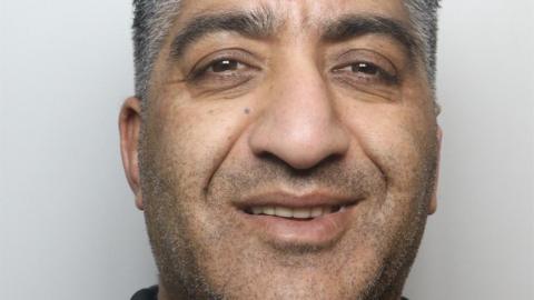 A police custody photo of a man with short grey and black hair and brown eyes smiles. A grey background behind him.